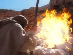 Person in robe and head covering holding a staff, facing a burning bush in rocky desert.